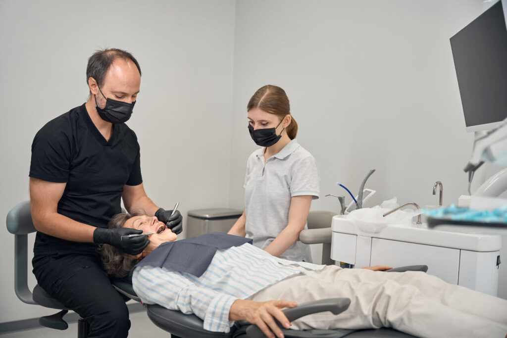 Middle-aged doctor treats an elderly lady's teeth, a young assistant helps him with his work